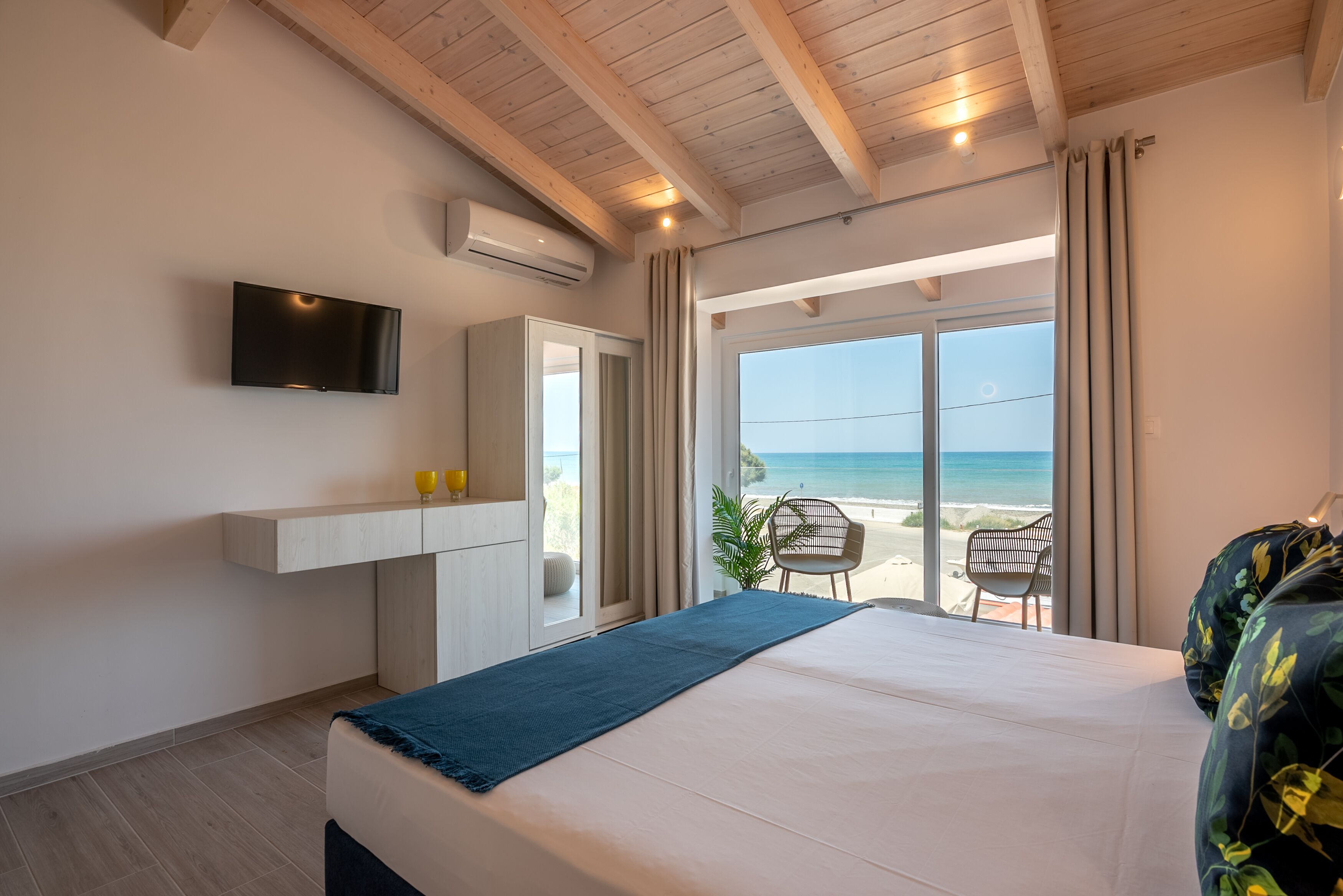 Ein Schlafzimmer mit Bett, einem an der Wand befestigten Fernseher, einem Fenster mit Vorhängen und Blick auf den Strand.