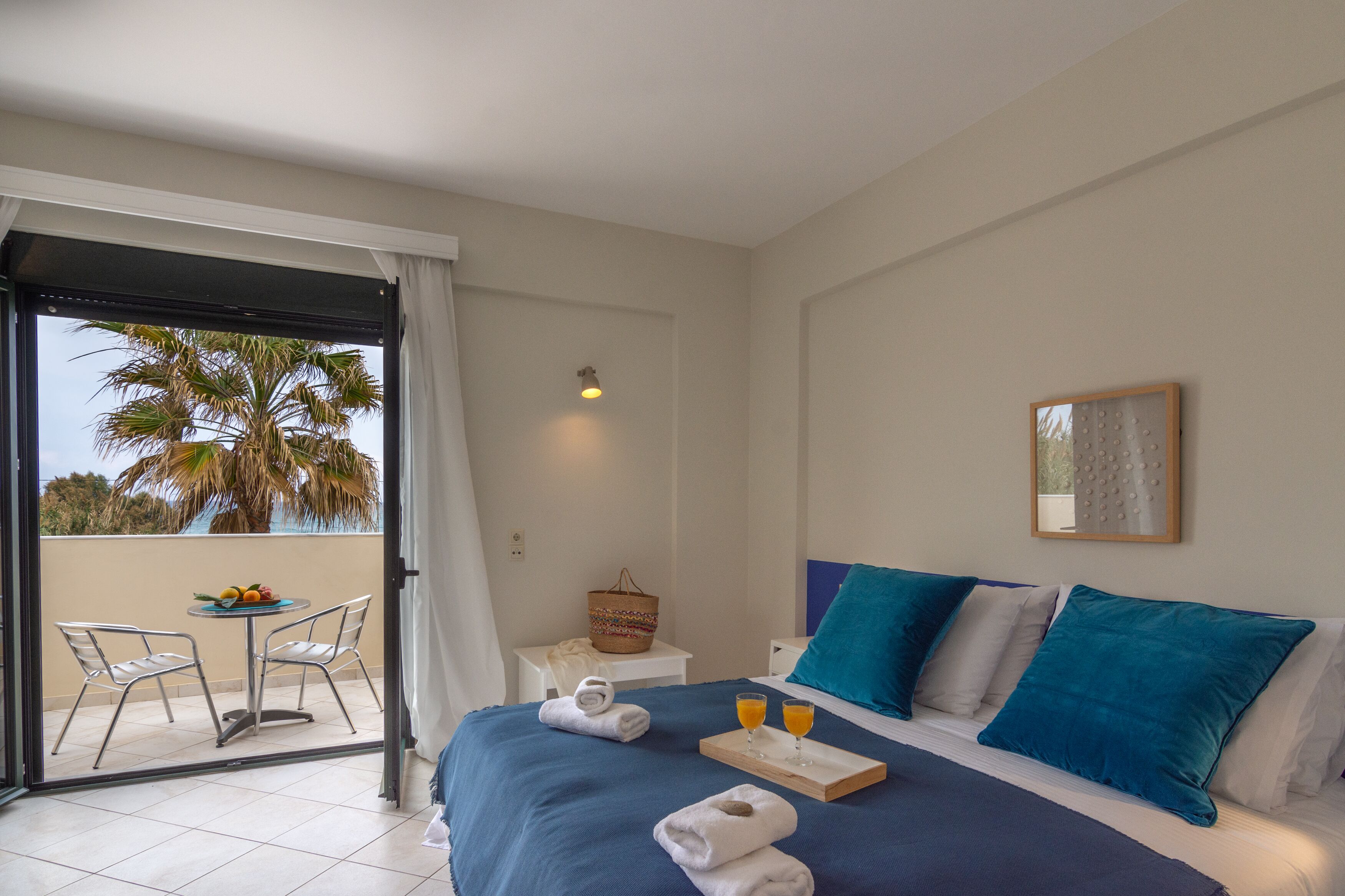 Habitación de hotel con cama, una mesita con frutas y bebidas, una silla y vista a palmeras a través de una puerta corrediza.