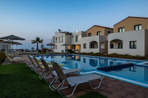 A swimming pool with lounge chairs, a building with a modern design, and a clear sky. - GT Beach Hotel (Platanias)