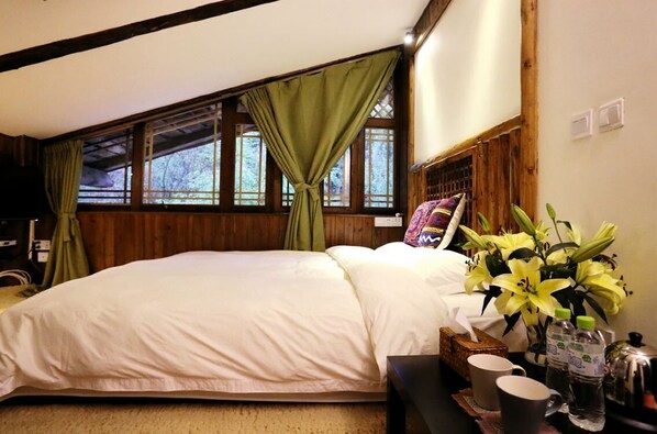 A bedroom with a large bed, wooden walls, a window with green curtains, and a bedside table with a vase of flowers and a water bottle. - Sunny Valley Inn (Deqin)