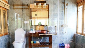 Un baño con inodoro, lavamanos con tocador de madera, ducha y zapatillas cerca de la puerta.