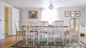 Uma sala de jantar com uma mesa de madeira, cadeiras brancas, um candelabro e um quadro na parede.