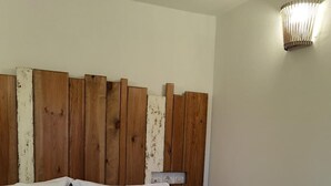 Una cama con sábanas blancas y una colcha de motivos florales, un cabecero de madera con acabado envejecido y una lámpara fijada a la pared.
