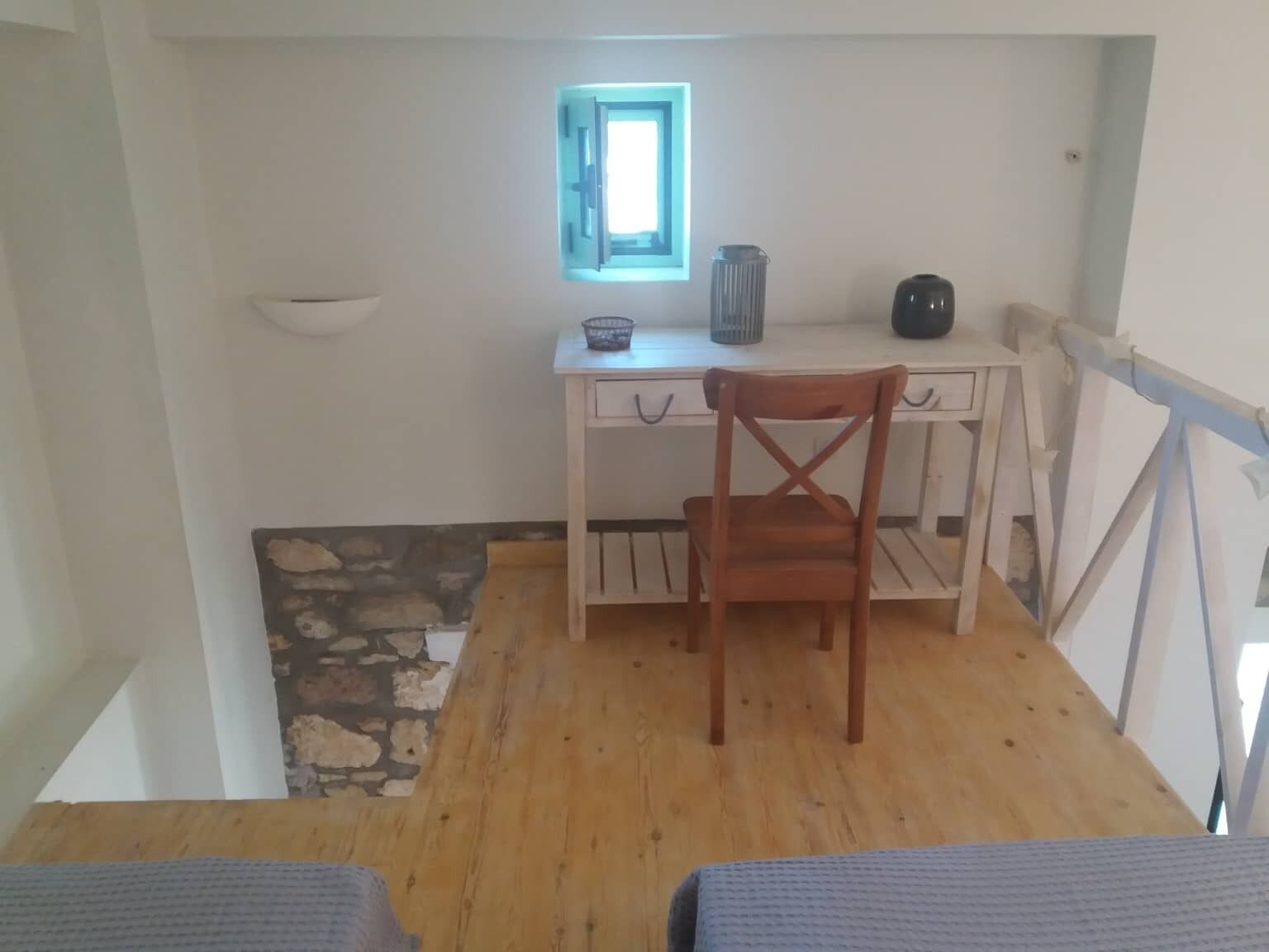 A small, bright room with a wooden desk, a chair, a small window, and a stone wall.