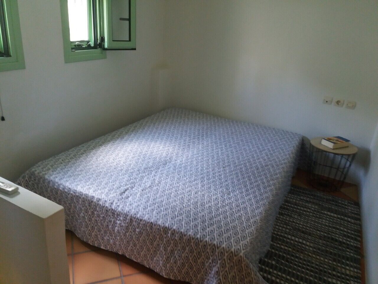 Una piccola camera con un letto singolo, una coperta a motivi decorativi, un tavolino con un libro e una finestra con cornici verdi.