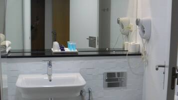 Un baño pequeño con un lavamanos blanco, un inodoro y una ducha. Se ve un espejo sobre el lavamanos, y un perchero en la pared.
