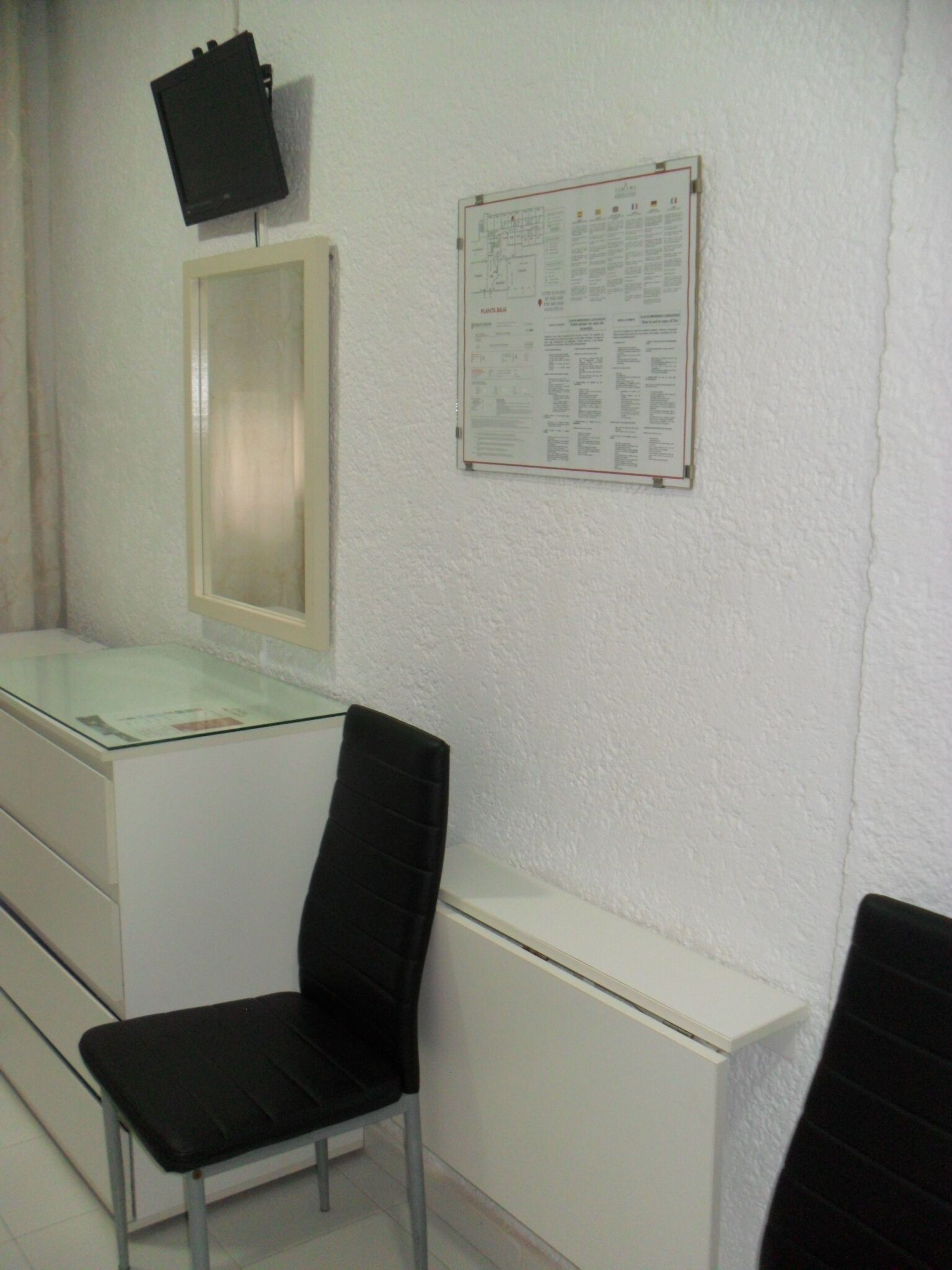 Een steriele ruimte met een bureau, een stoel, een wandtelevisie en een prikbord.