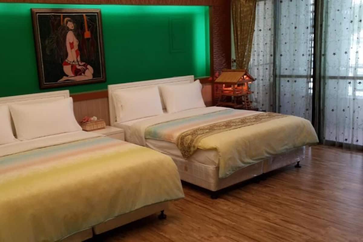 A double bed with a rainbow-colored blanket, a green wall with a framed artwork, and a wooden nightstand with a lamp.
