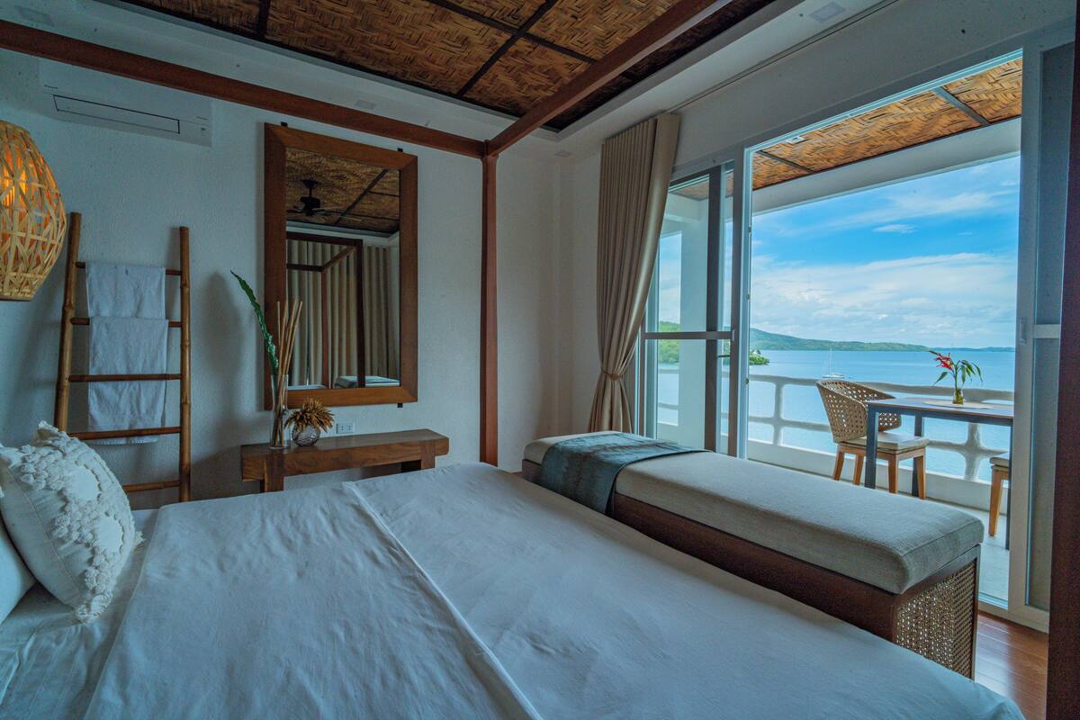 A bedroom with a large bed, a wooden bench, a small table, and a view of the sea through a balcony.