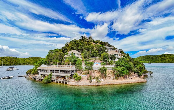 Une île tropicale avec une grande maison entourée d’une végétation luxuriante et sous un ciel bleu azur.