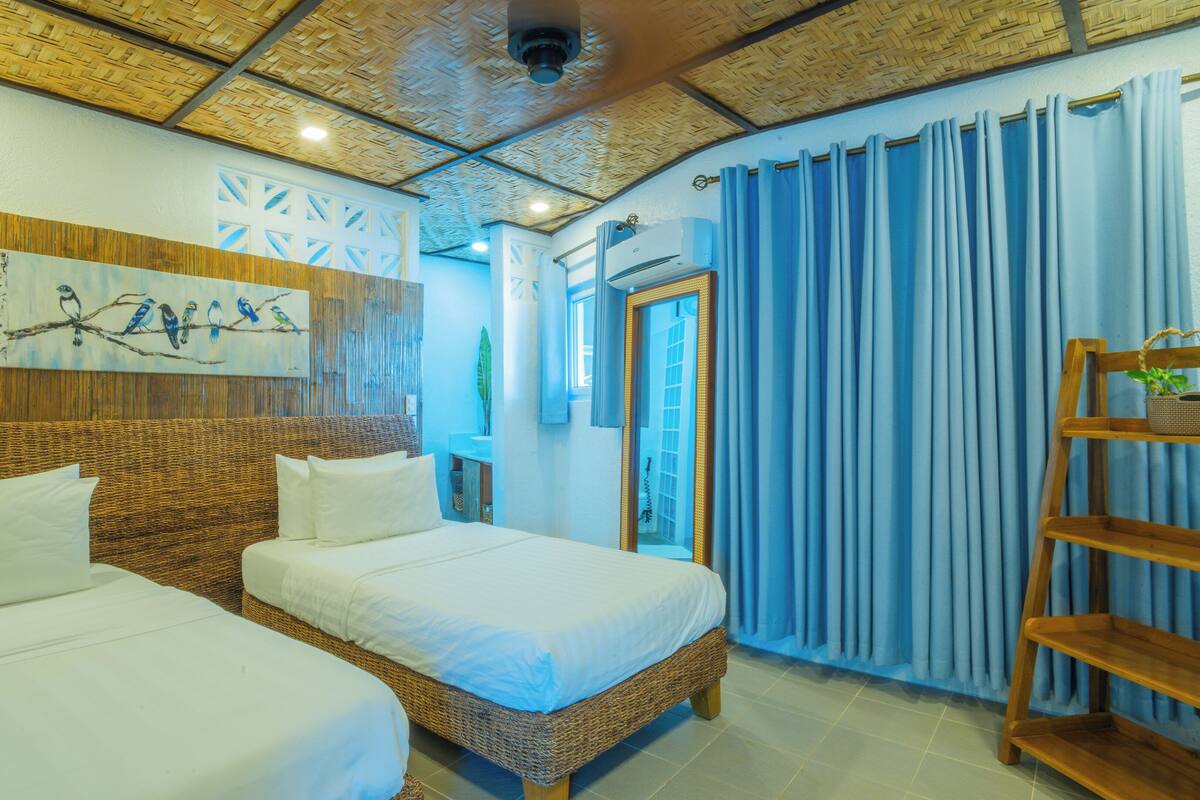 A room with two beds, a wooden headboard, a woven wall panel, a ceiling with a woven pattern, a window with blue curtains, and a wooden shelf with a plant.