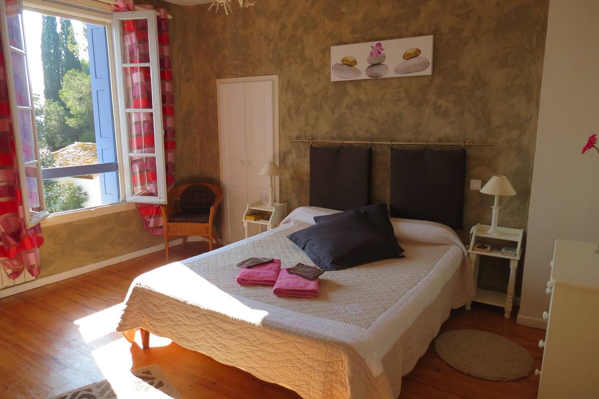A bedroom with a large bed, a chair, a nightstand, a lamp, and a window with curtains.