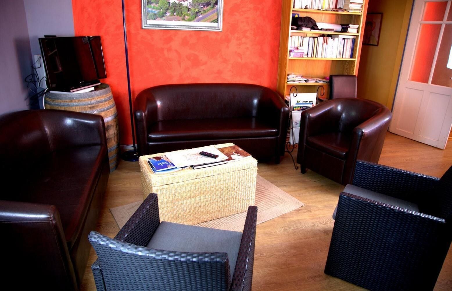 Ein Wohnzimmer mit einem dunkelbraunen Ledersofa, zwei Korbsesseln, einem kleinen Couchtisch mit Büchern und einer Fernbedienung, einem Bücherregal und einem Fernseher auf einem Holzboden.