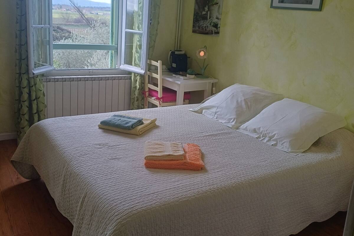 A neatly made bed with white linens, a small table with a lamp, and a window with curtains.