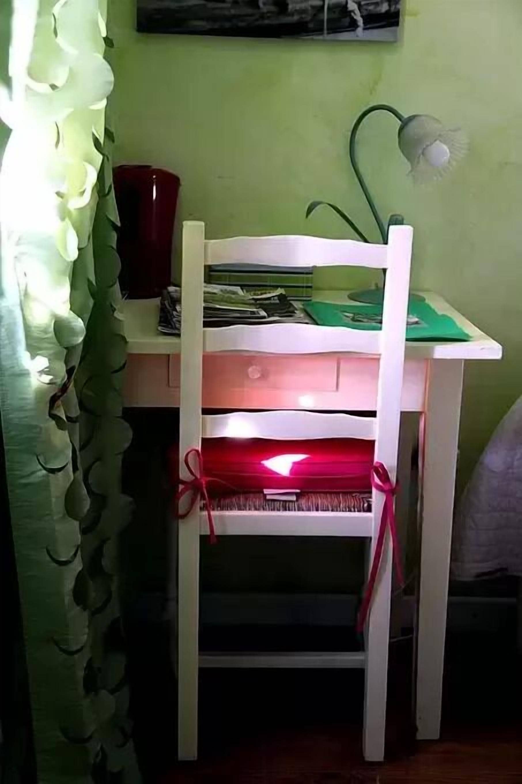 Une chaise blanche ornée d’un ruban rouge noué autour de l’assise, placée devant un bureau sur lequel se trouvent des papiers et un classeur vert.