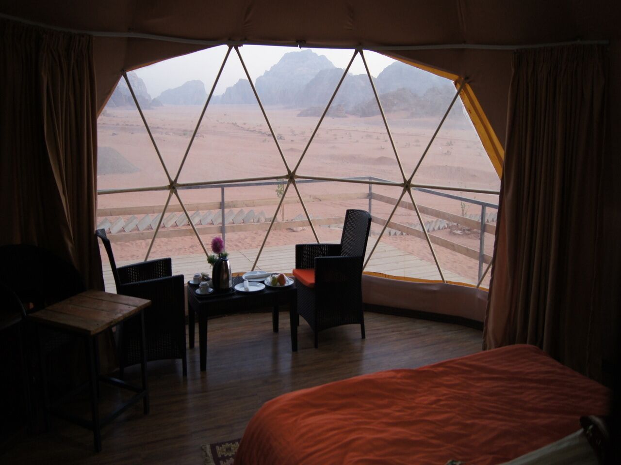 A room with a large window offering a view of a desert landscape. There is a bed, two chairs, a small table with a vase and a plate.