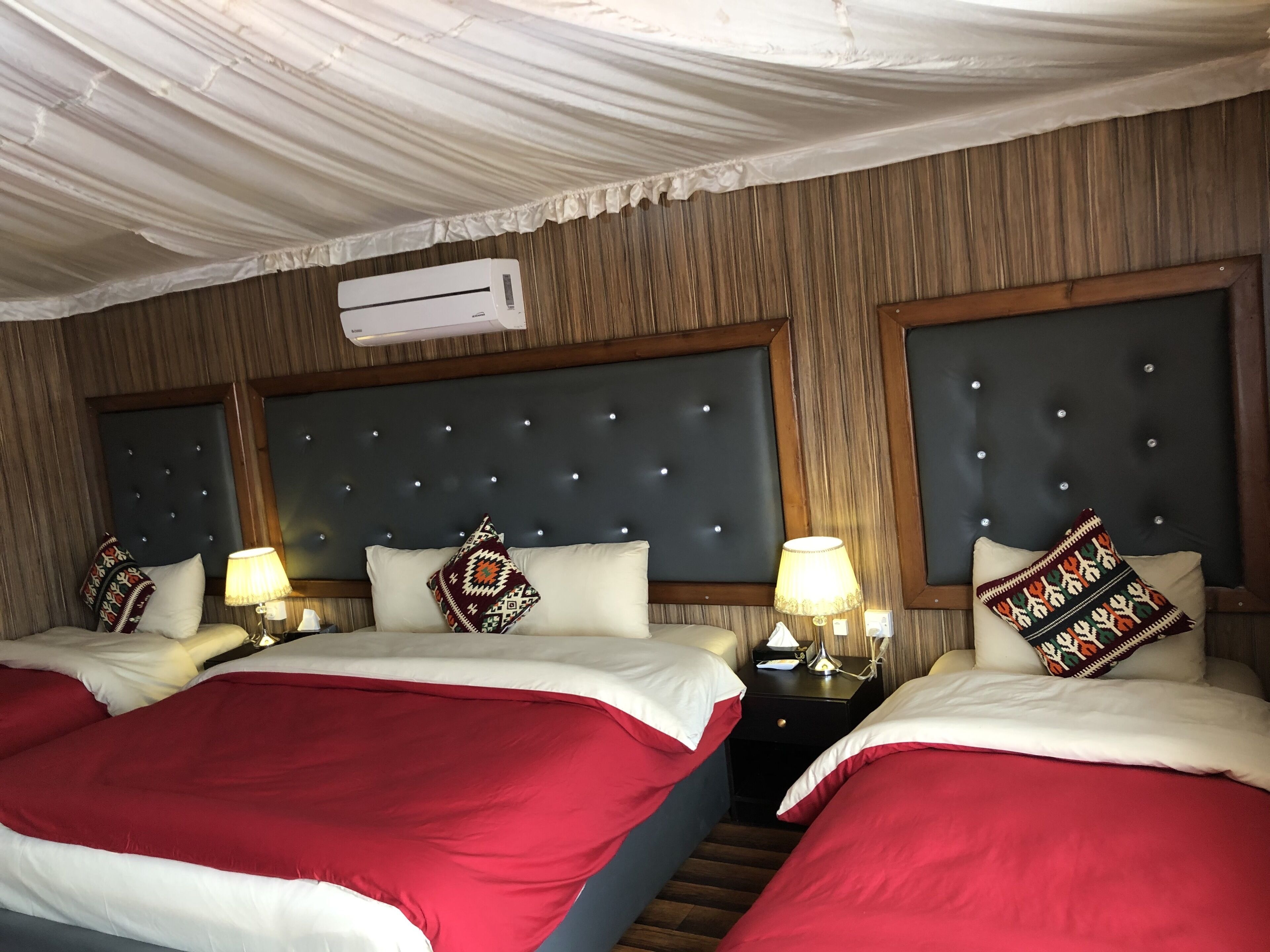 Une chambre d’hôtel avec deux lits, chacun recouvert d’une couvre-lit rouge et blanche, dotée d’une tête de lit sombre à motifs capitonnés et d’un climatiseur mural.