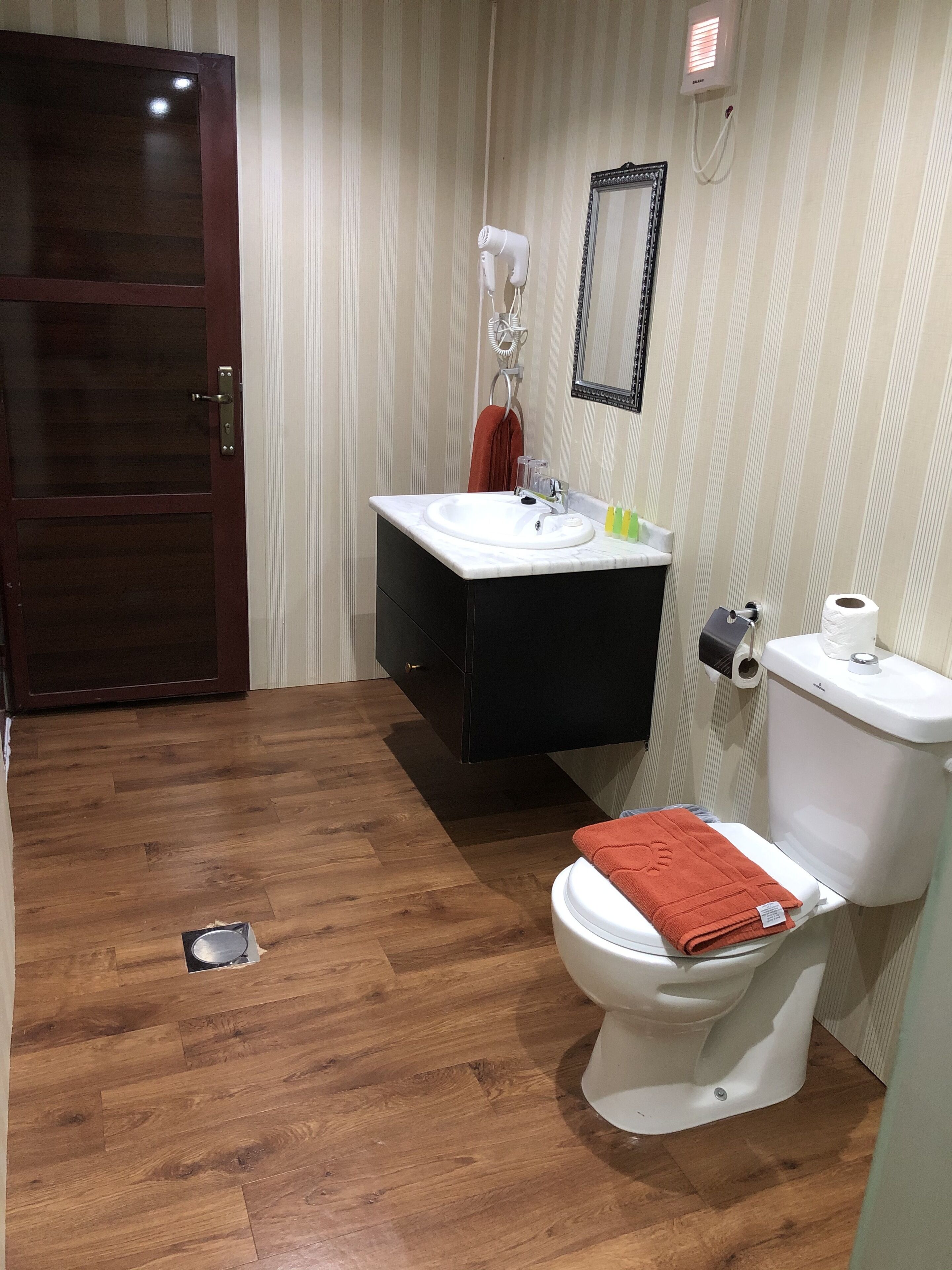 Een badkamer met een houten vloer, een wit toilet, een zwart kastje, een witte wastafel, een spiegel en een handdoekhouder.