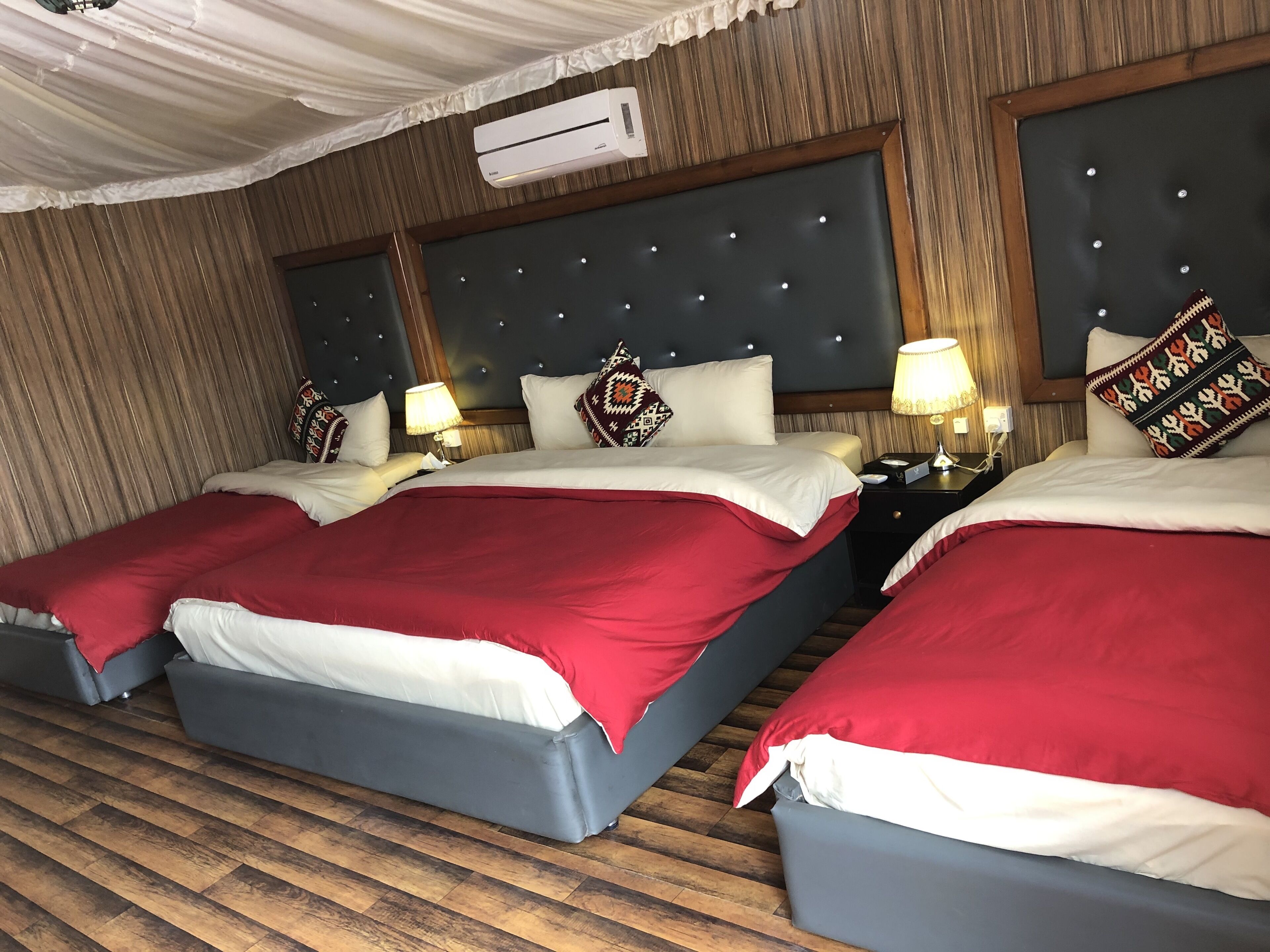 A hotel room with three beds, each with a red and white bedspread, wooden headboards, and patterned pillows. There is an air conditioning unit on the wall, and the room has wooden flooring.