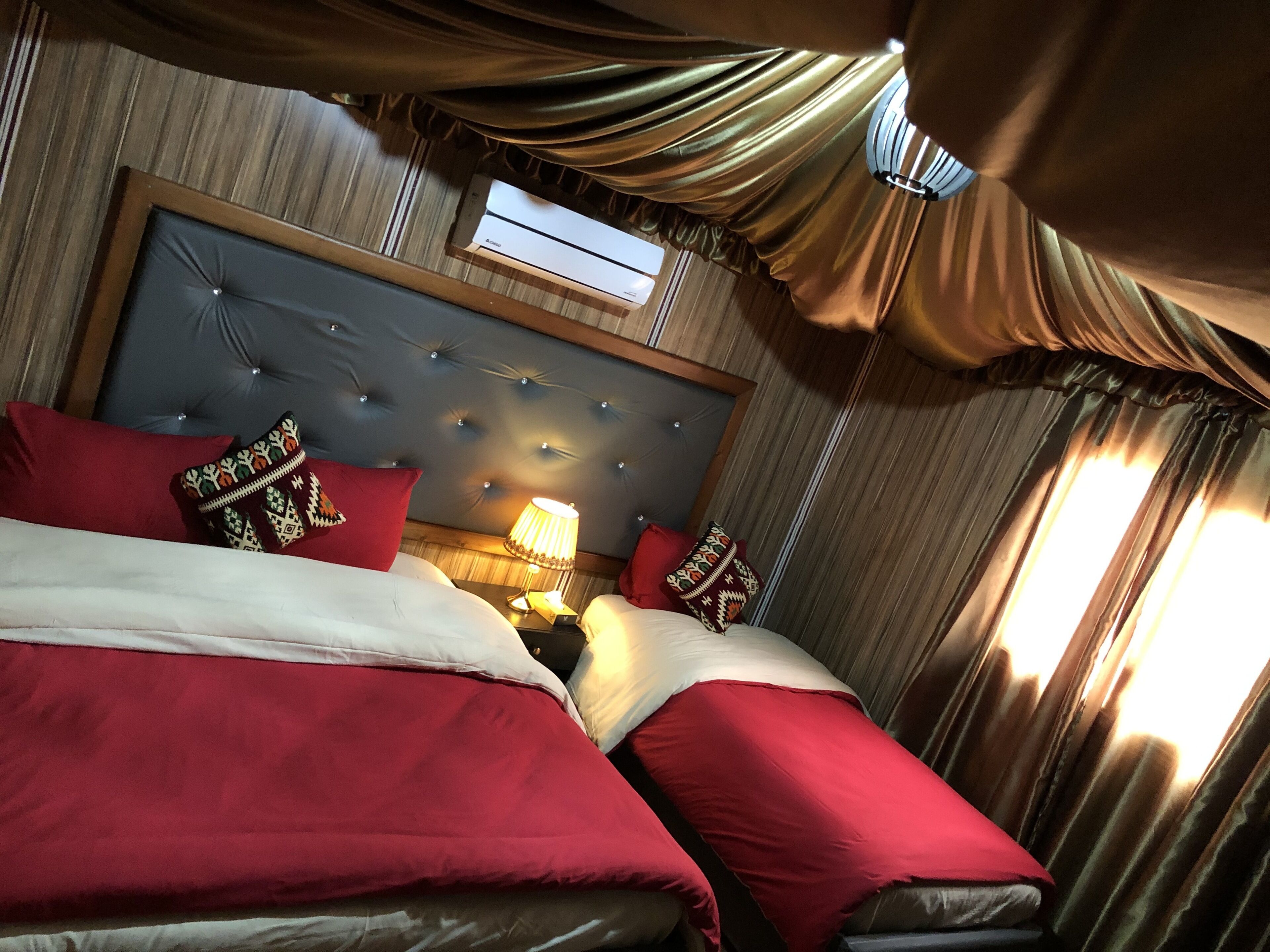 A hotel room with a large bed, red bedding, a tufted headboard, a bedside lamp, and a window with curtains.