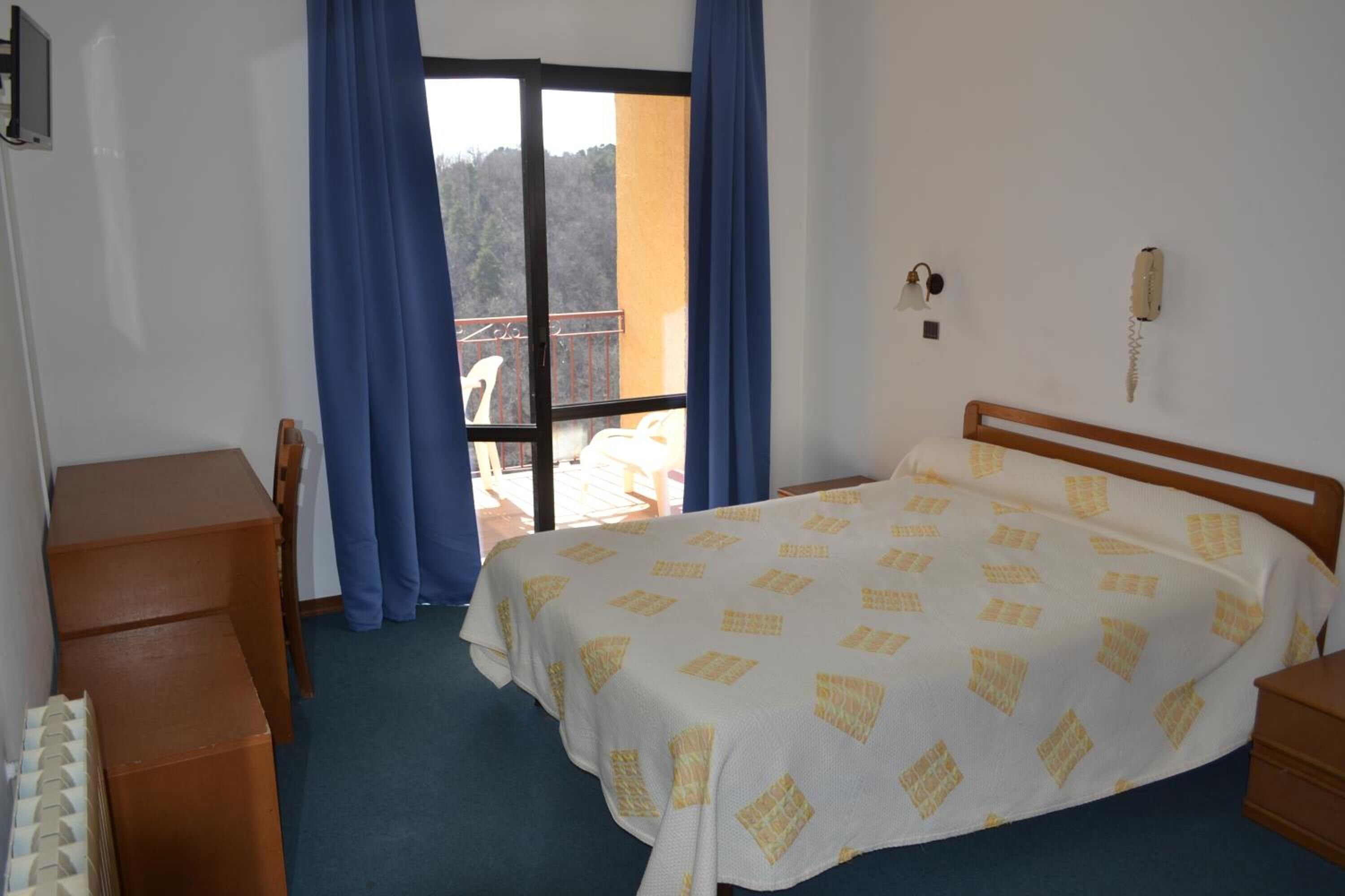 A hotel room with a bed, a wooden nightstand, a desk, and a chair. There is a balcony with blue curtains and a view of trees.