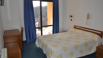 A hotel room with a bed, a wooden nightstand, a desk, and a chair. There is a balcony with blue curtains and a view of trees.