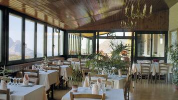 Een restaurant met houten plafond en grote ramen, tafels gedekt met witte tafelkleden en stoelen, en potplanten.