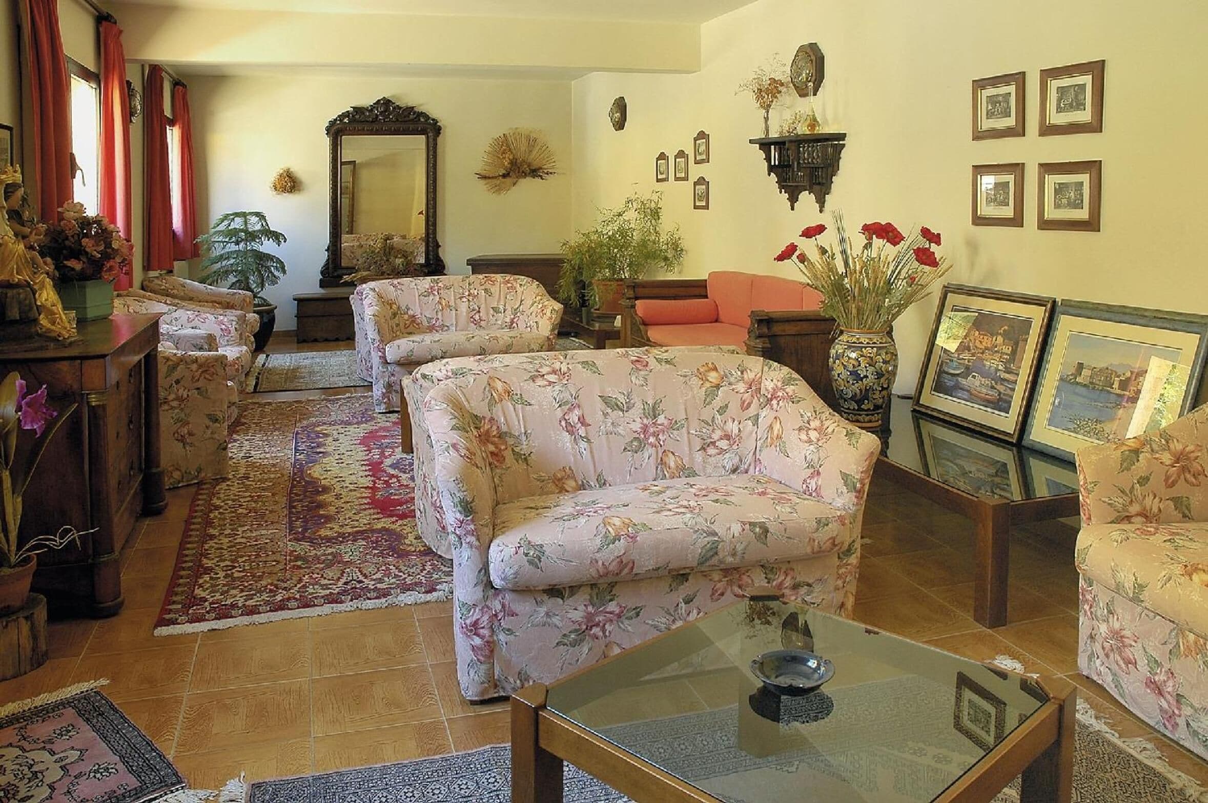 A living room with floral-patterned sofas, a glass coffee table, and a variety of decorative items including a mirror, plants, and framed pictures on the walls.