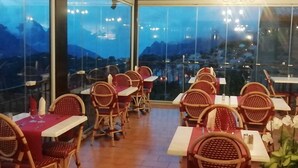 A restaurant with wicker chairs and tables, large windows offering a view of the city and mountains. - Hotel Aitone (Evisa)