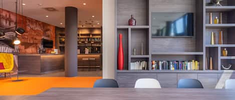 Uma sala de conferências moderna com uma mesa de madeira, estantes cinzentas com livros e um vaso vermelho.