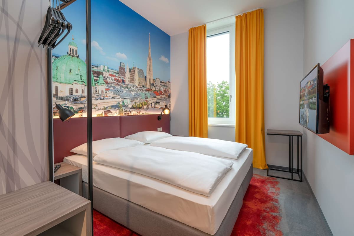 A hotel room with a large bed, a desk, a chair, a TV, and a window with curtains.