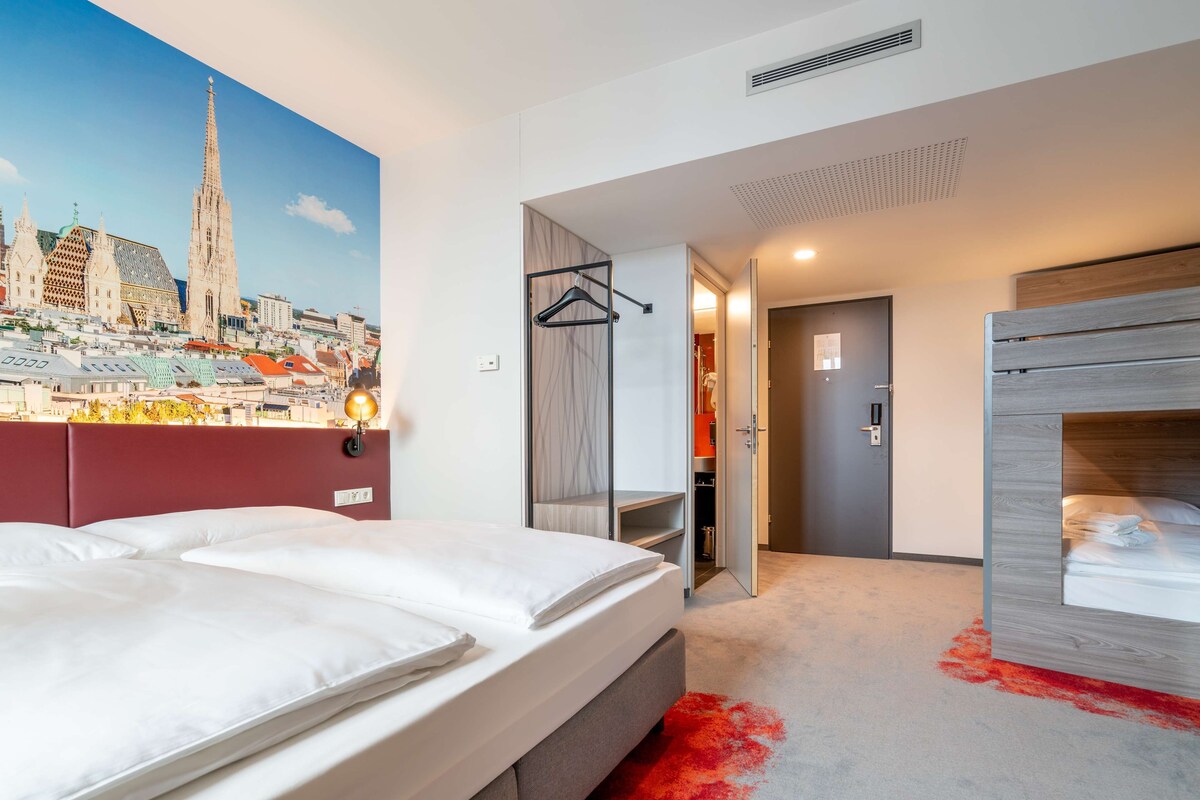 A hotel room with a large bed, a wardrobe, a mirror, and a view of a cityscape mural.