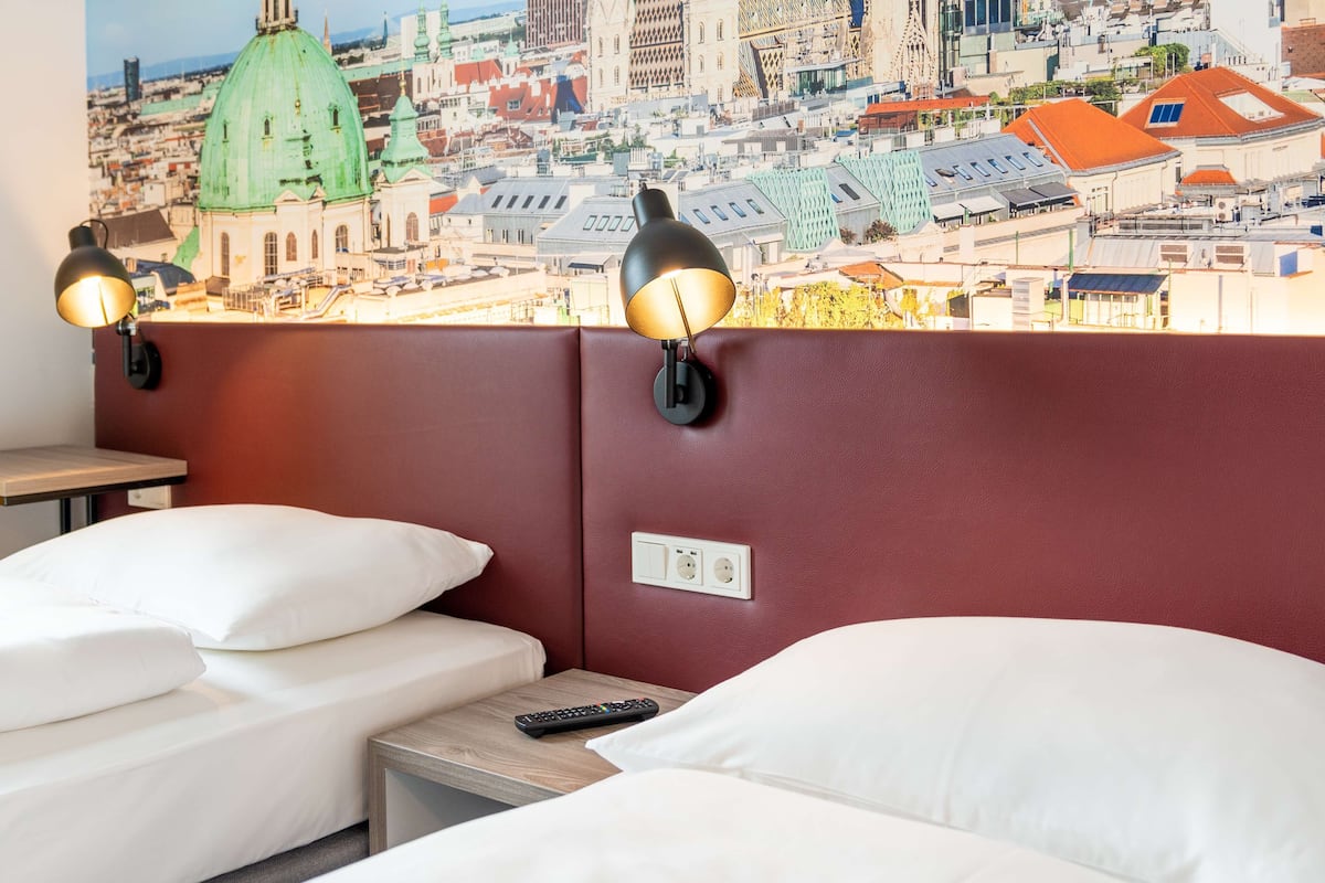 A hotel room with two beds, a cityscape mural on the wall, and two wall-mounted lamps.