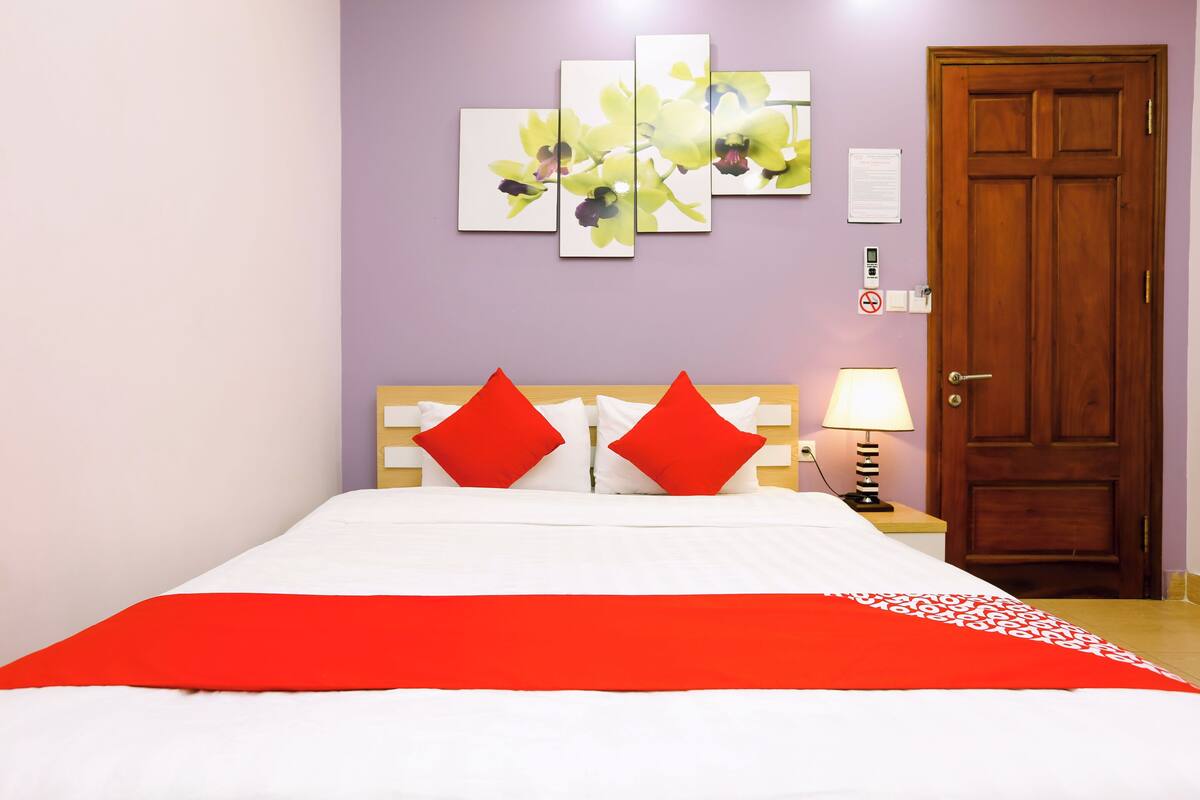 Een netjes opgemaakt bed met een rood-witte spreidek, twee rode kussens en een houten hoofdbord. Daarboven hangt een kunstwerk van gele bloemen aan de muur.