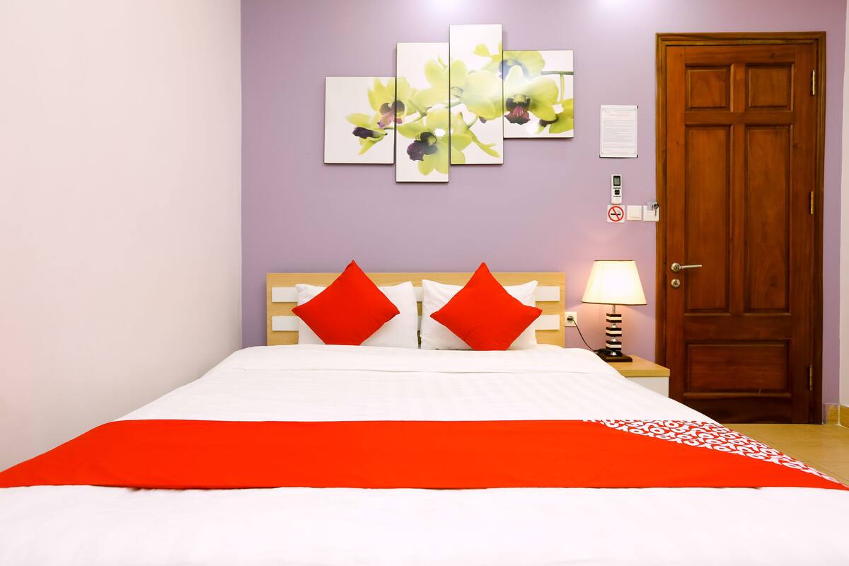 Een netjes opgemaakt bed met een rood-witte spreidek, twee rode kussens en een houten hoofdbord. Daarboven hangt een kunstwerk van gele bloemen aan de muur.