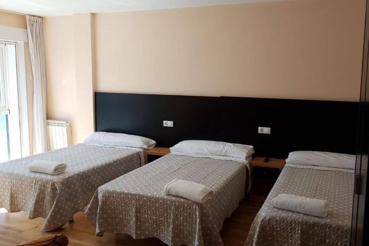 A hotel room with three single beds, each with a white pillow and a folded towel. The room has a wooden floor and a large window with curtains.