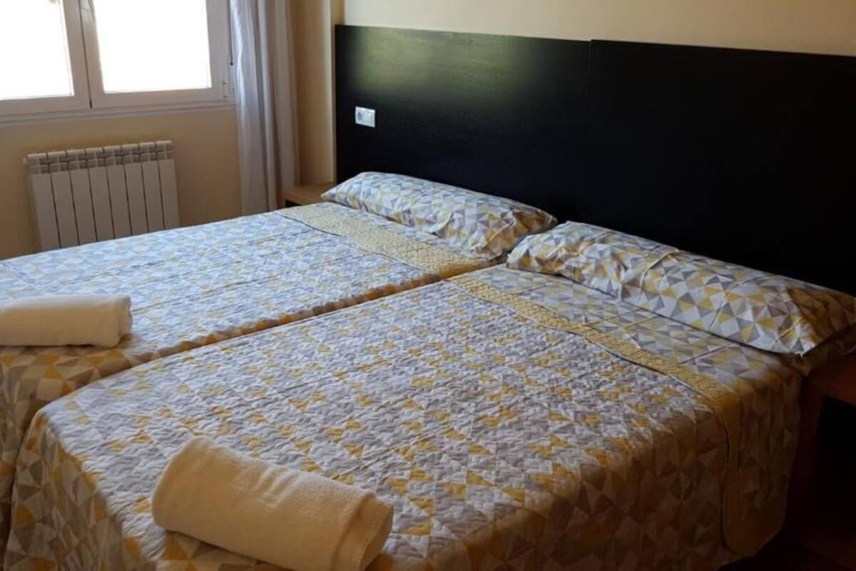 A room with two beds, each with a patterned bedspread and pillows, a radiator, and a window with curtains.