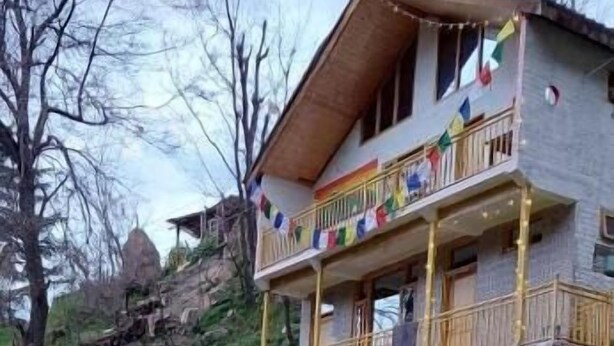 Un edificio de dos pisos con un balcón, una persona sentada sobre un tocón de árbol y una tienda de campaña verde cerca.