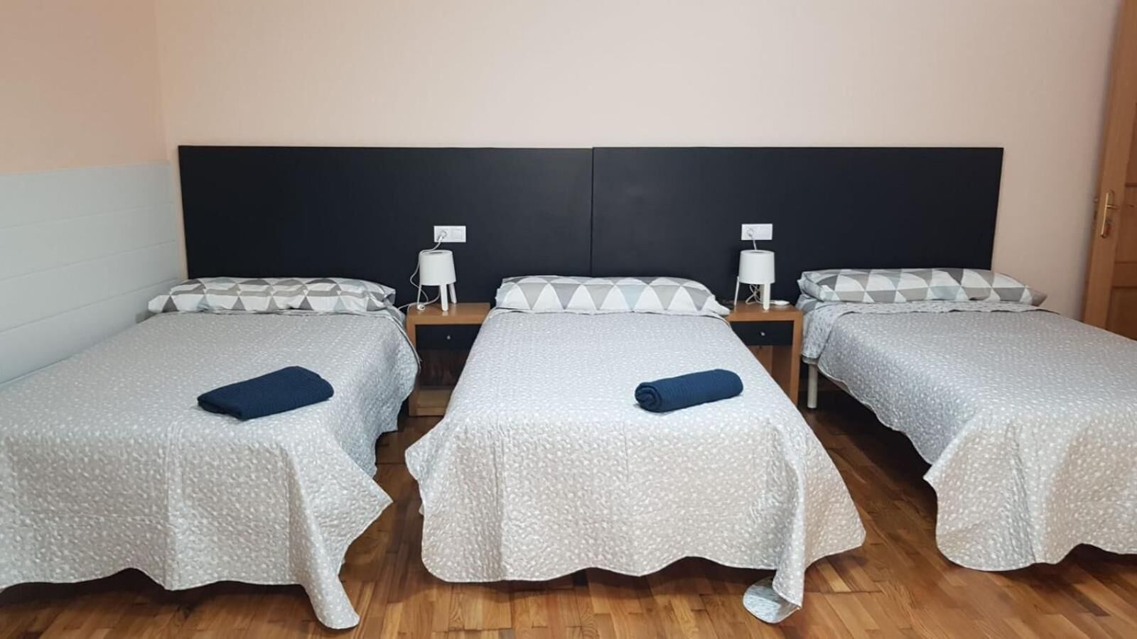 A room with three single beds, each with a white textured bedspread and a blue towel. There are two bedside tables with lamps and a wall socket above each bed. The room has wooden flooring and a dark headboard behind the beds.