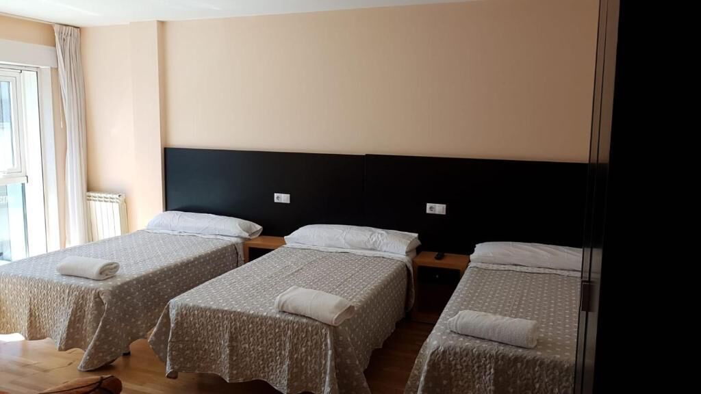 Ein Hotelzimmer mit drei Einzelbetten, jedes mit einem weißen Kopfkissen und einem gefalteten Handtuch. Der Raum hat einen Holzboden und ein großes Fenster mit Vorhängen.