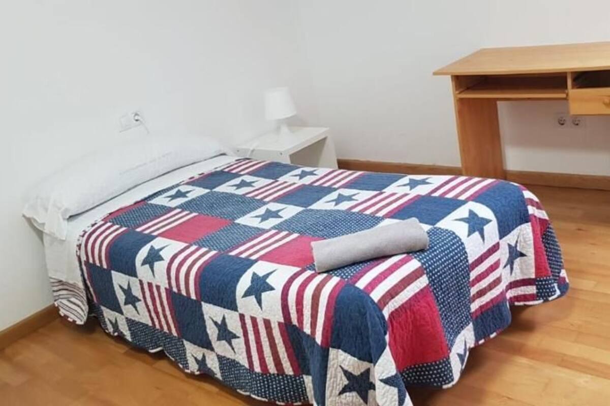 Un lit simple avec une couette, une table de chevet blanche, un petit bureau et un plancher en bois.