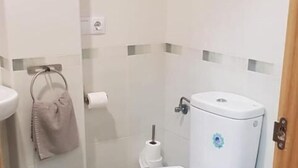 Un baño pequeño y limpio con un inodoro blanco, un cubo de basura azul y un lavamanos blanco.