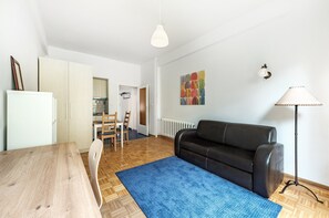 A modern living room with a wooden dining table, a black leather sofa, a blue rug, and a white pendant light.