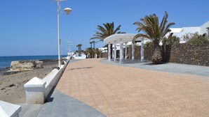 Une promenade en bord de mer, bordée d’un mur de pierres, de palmiers et d’un bâtiment blanc surmonté d’une pergola.