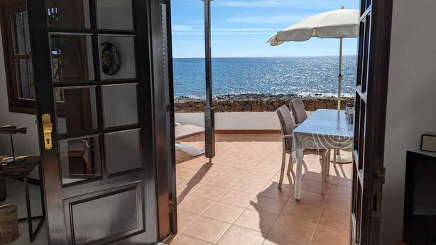 Une porte ouverte donnant sur un patio équipé d’une table et de chaises, d’un parasol blanc et offrant une vue sur la mer.