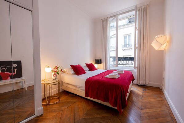 A hotel room with a bed, a bedside table with a lamp, a window with curtains, and a view of the outside balcony.