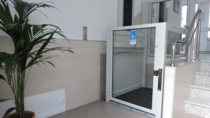 A modern elevator with a glass door, a potted plant, and a blue accessibility sign. - Hostal Delia (Álora)