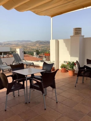 An outdoor patio with a table and chairs, a view of the mountains, and a white building in the background. - Hostal Delia (Álora)