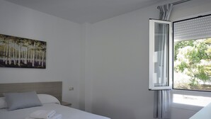 A hotel room with a bed, a painting, a window with blinds, and a desk with a remote control. - Hostal Delia (Álora)