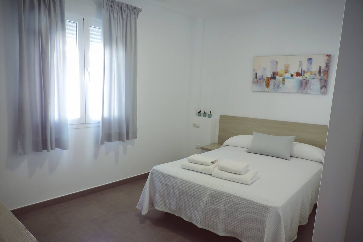 A hotel room with a bed, a nightstand, a window with curtains, and a painting on the wall.